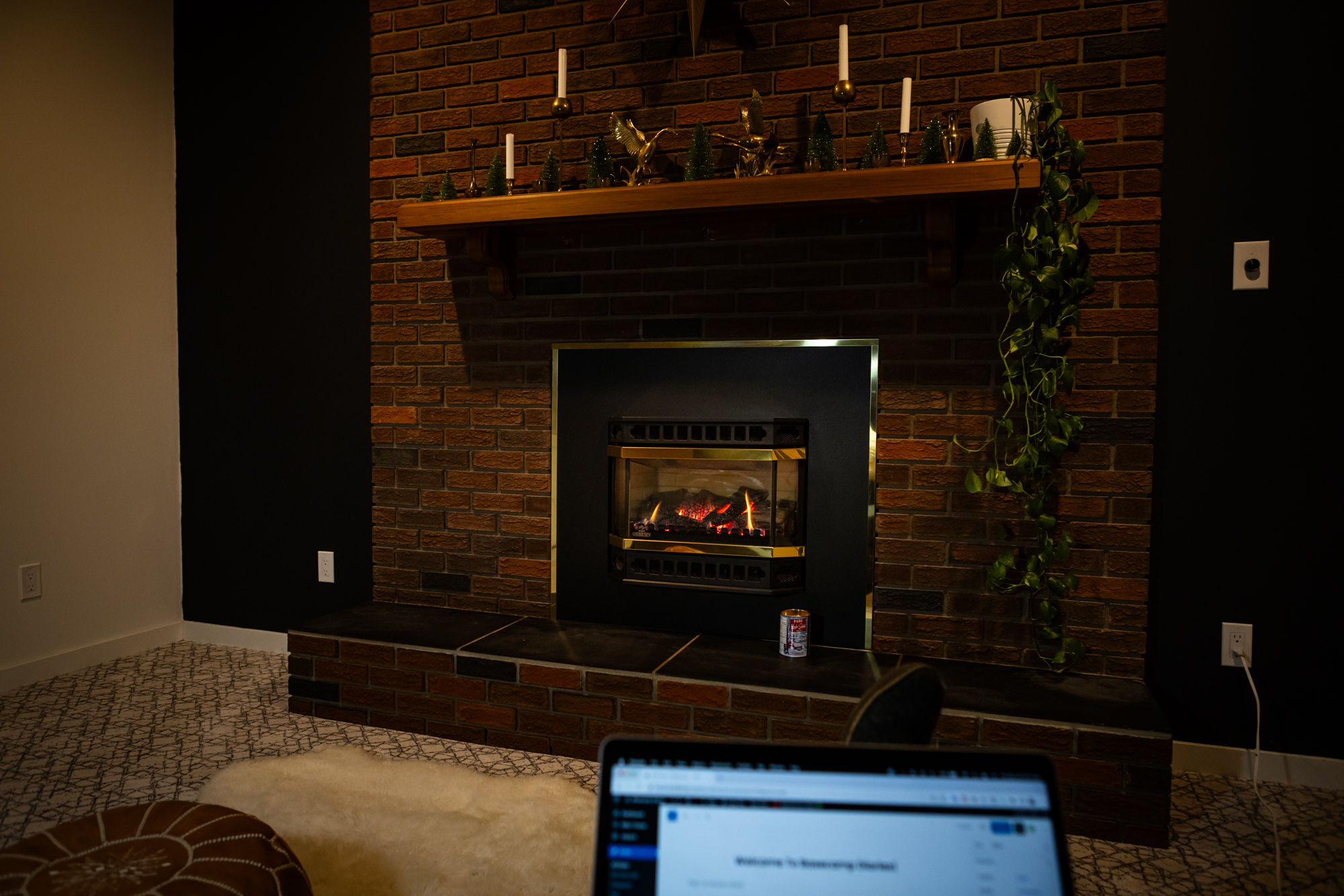 POV sitting on a sofa with legs out, laptop on lap in front of a fireplace. There is a sheepskin rug on the floor. Mantel is decorated in brass candleholders, brass Canada geese figurines, and green bottle brush trees, a pothos is to the right