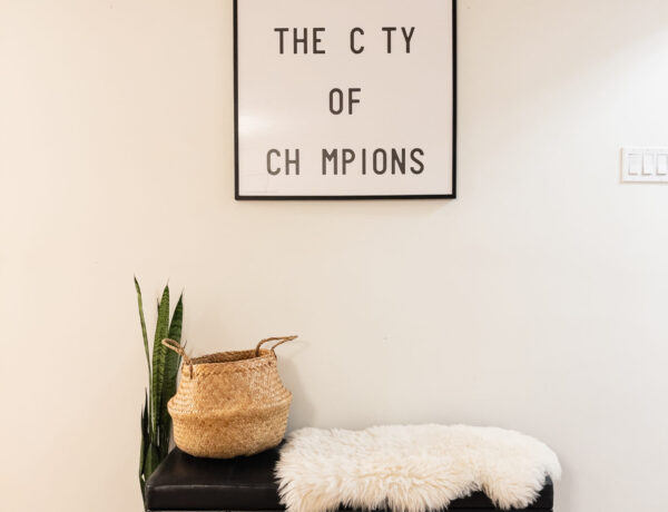 Black leather bench, with a seagrass basket white sheepskin rug. Next to the bench is a snake plant, and a small potted hoya. Framed art on the wall reads "Welcome to the city of champions" with a few letters missing (E in welcome, I in city, A in Champions)