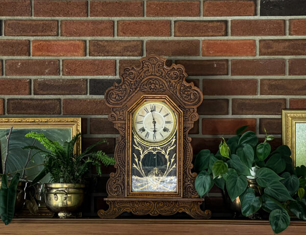 Antique Waterbury mantle clock sits on a wood mantle with a brick wall. Fern in a brass pot to the left, and a heart leaf philodendron to the right. Gold framed mirror in the background